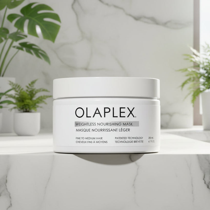 A jar of Olaplex Weightless Nourishing Mask displayed on a white marble surface, surrounded by small potted green plants in soft natural daylight.